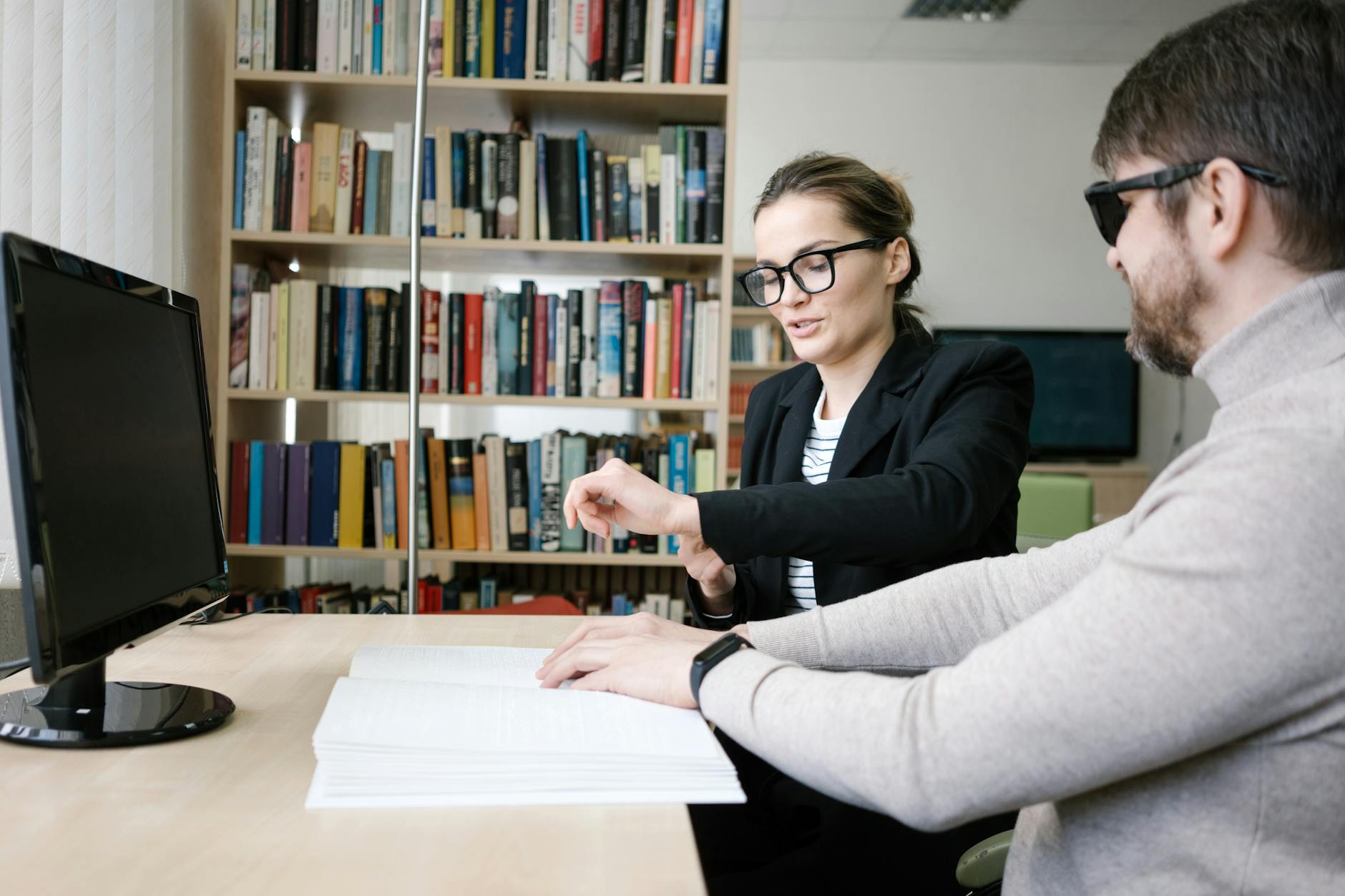 Kobieta pomaga niewidomemu mężczyźnie czytać Braille’a przy biurku w bibliotece